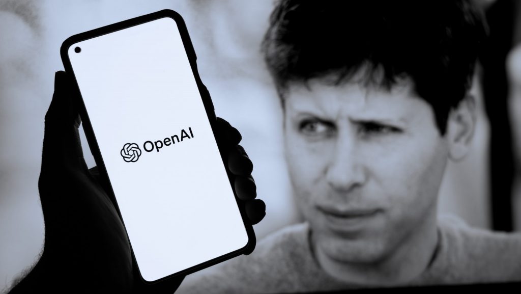 Pessoa segurando celular com logomarca da OpenAI na tela na frente de monitor exibindo foto do CEO da empresa, Sam Altman