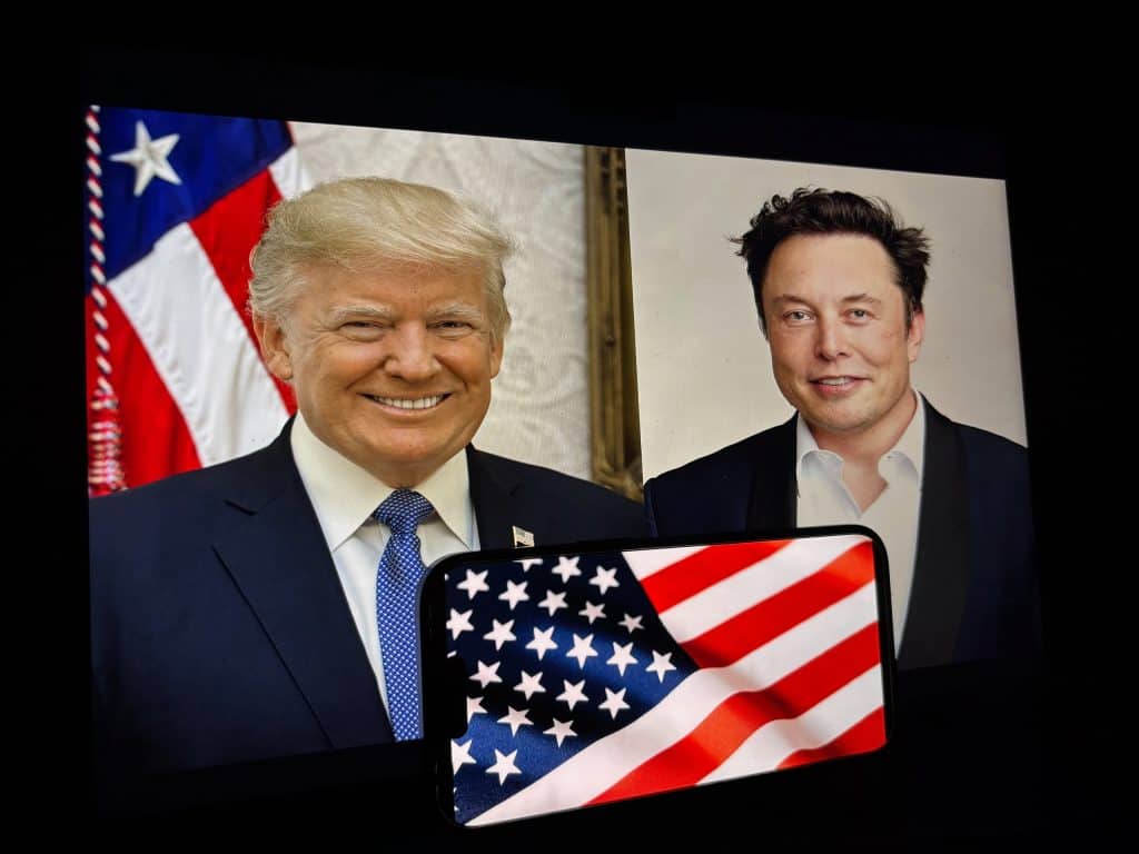 Ao fundo, fotos de ELon Musk (direita) e Donald Trump (esquerda); à frente, parte da bandeira dos EUA tremulando na tela de um smartphone