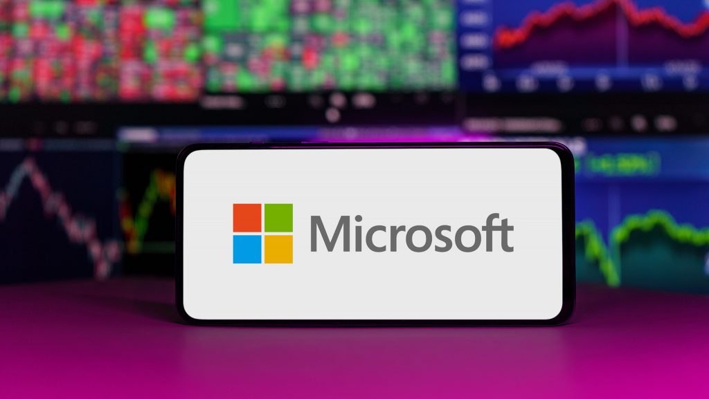 Logo da Microsoft em um smartphone; atrás, telas com gráficos com ações