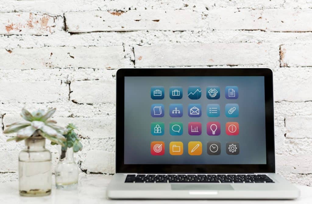 laptop aberto com diversos ícones de aplicativos na tela, em uma mesa branca e uma parede de tijolos brancos ao fundo