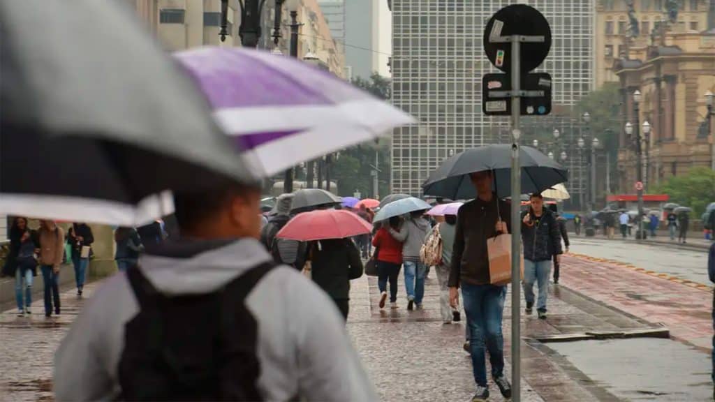 Pessoas agasalhadas e carregando guarda-chuvas andando em avenida de São Paulo