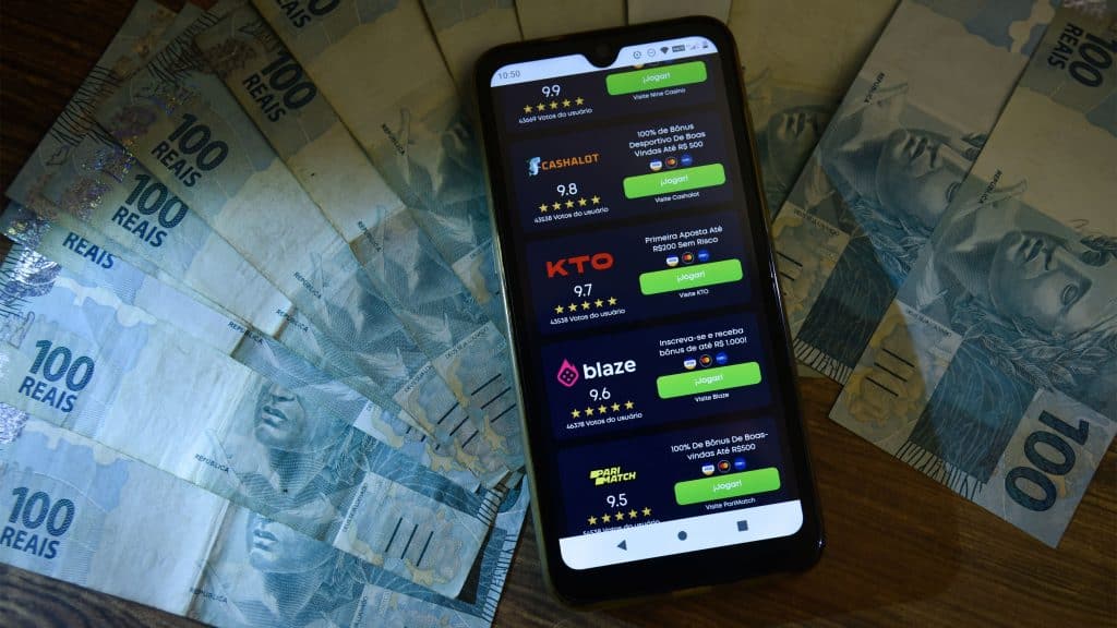 Celular com lista de bets que funcionam no Brasil em cima de notas de 100 reais
