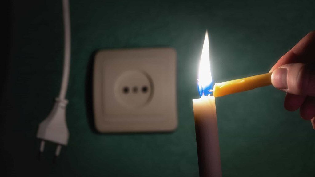 Ao fundo, um plugue de tomada desconectado e uma tomada ao lado; à frente, em foco, uma vela sendo acesa