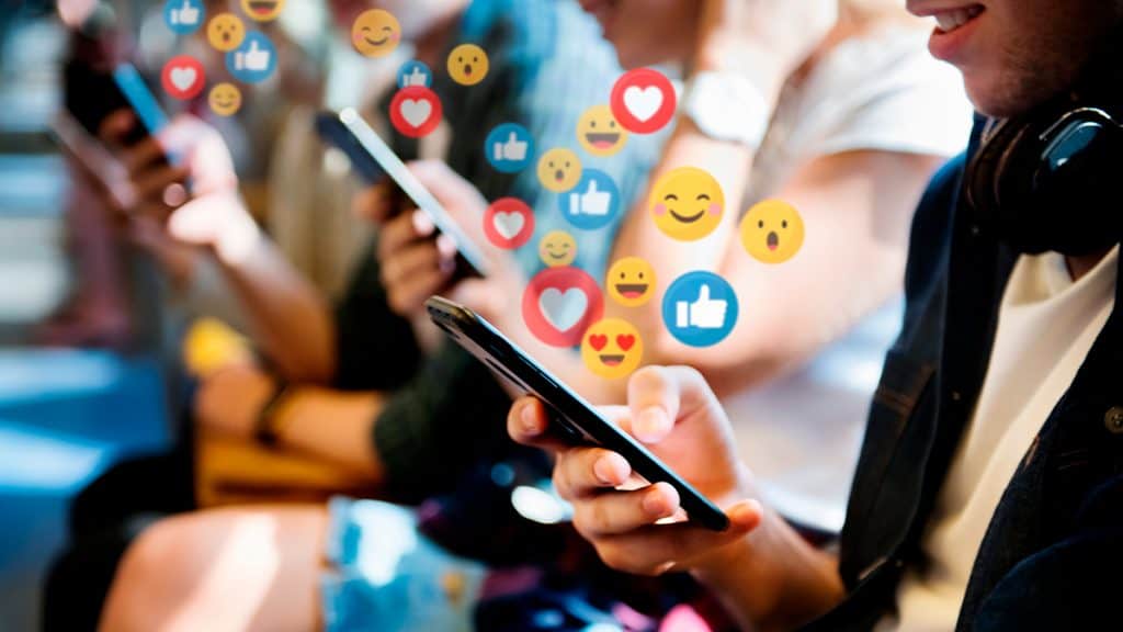 Jovens usando celular com emojis de reações de redes sociais saindo dos aparelhos