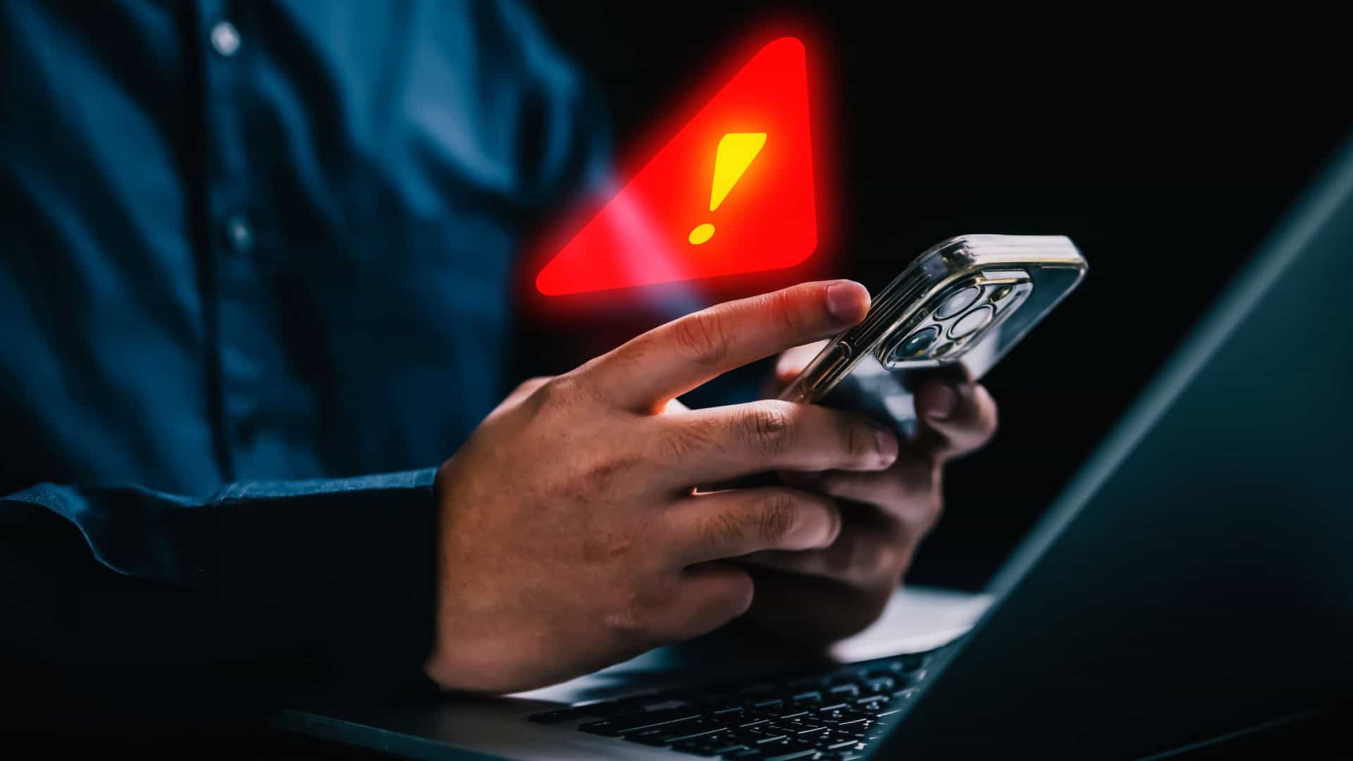 Pessoa usando iPhone em frente notebook com grande sinal de alerta holográfico em cima do celular