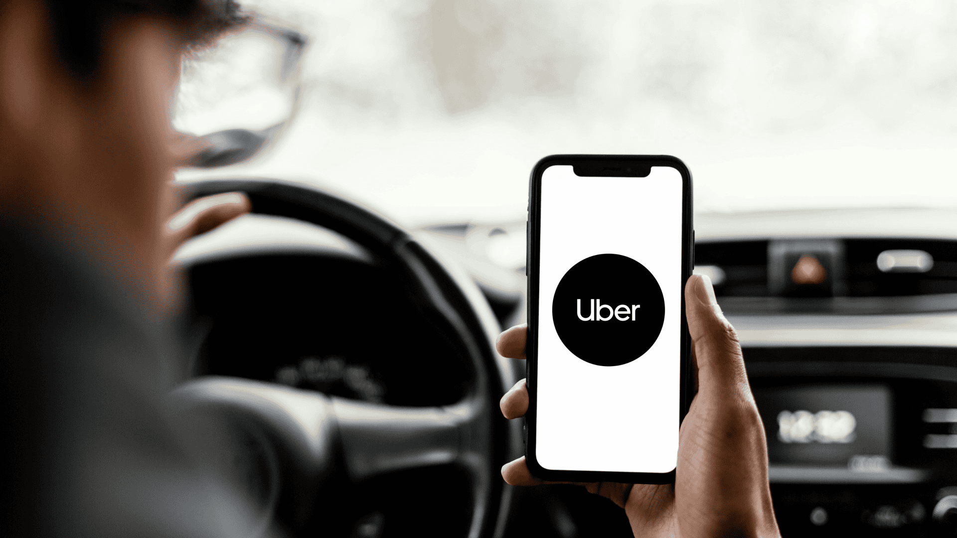 Motorista segura celular com o app da Uber aberto enquanto está ao volante, com interior do carro desfocado ao fundo