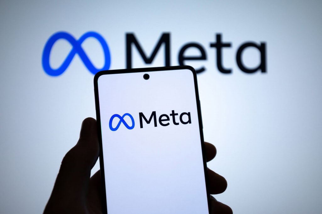 Logo da Meta em um smartphone e ao fundo, em uma tela grande