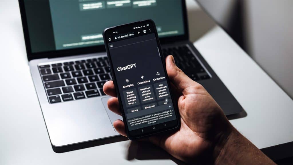 Pessoa segurando celular com ChatGPT aberto em frente a um MacBook