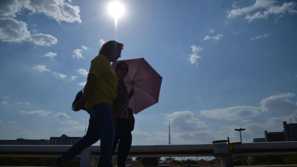 Duas mulheres andando na rua. Uma delas está com uma sombrinha rosa sobre a cabeça. O dia está ensolarado e parece quente.