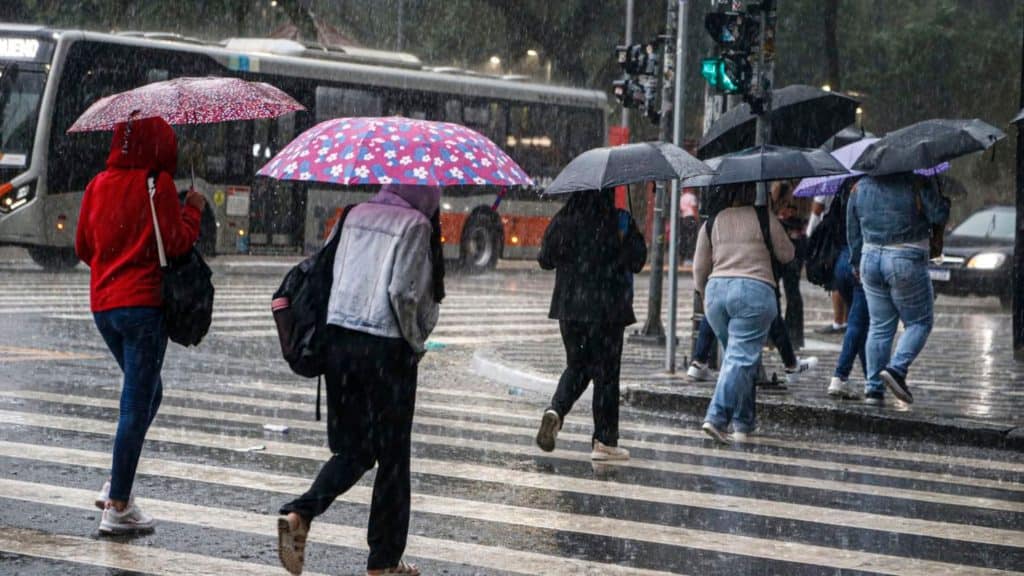 Pessoas atravessando avenida segurando guarda-chuva em dia chuvoso