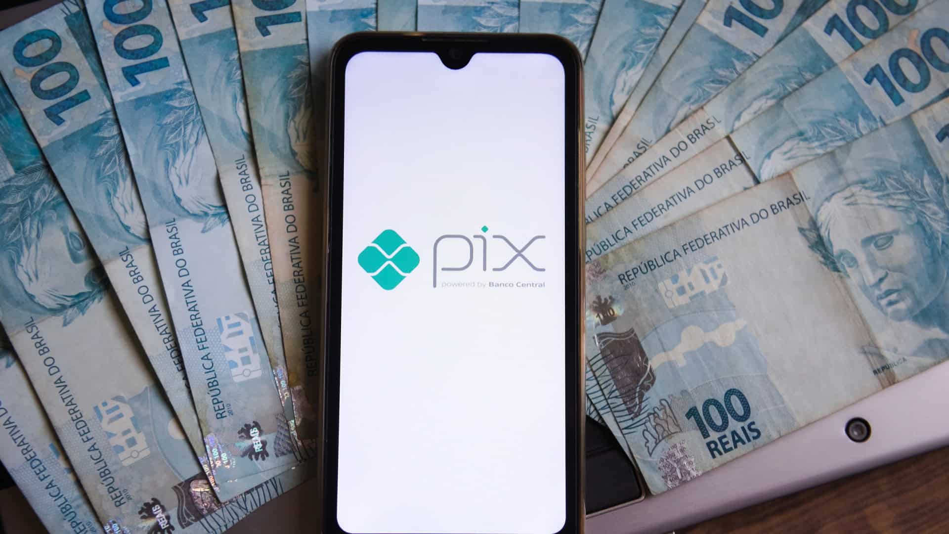 Notas de R$ 100 espalhadas; por cima, um smartphone com o logotipo do Pix